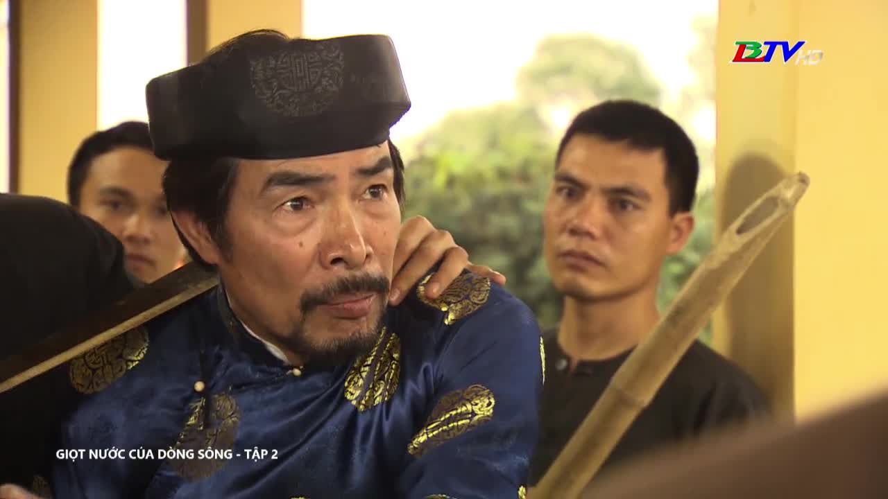 The man in the dark hat and blue tunic looks concerned, his face etched with worry. A younger man stands behind him, holding a bamboo pole and watching intently.
