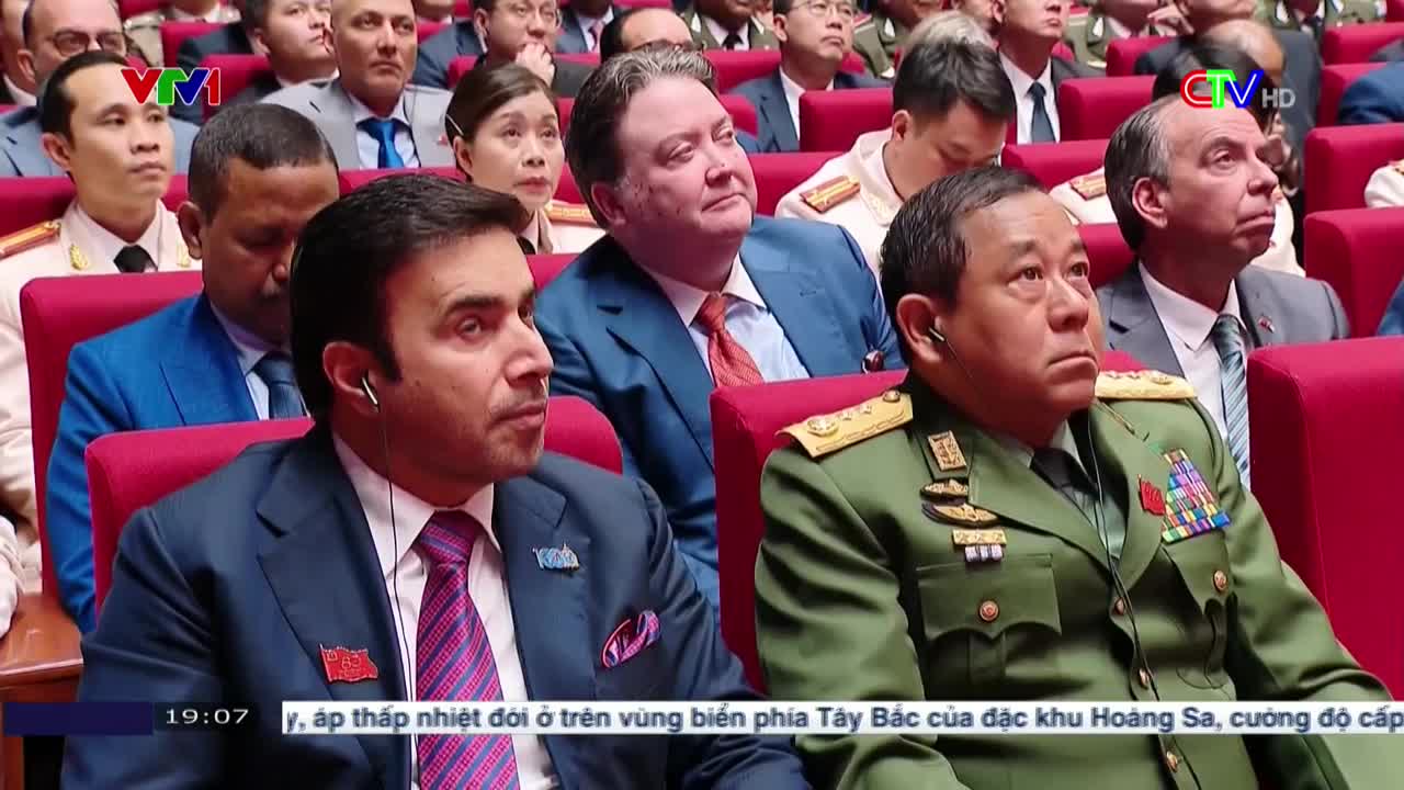 Men in suits and military uniforms sit in rows of red chairs, all looking in the same direction. The broadcast from Bac Lieu TV is showing a news report.
