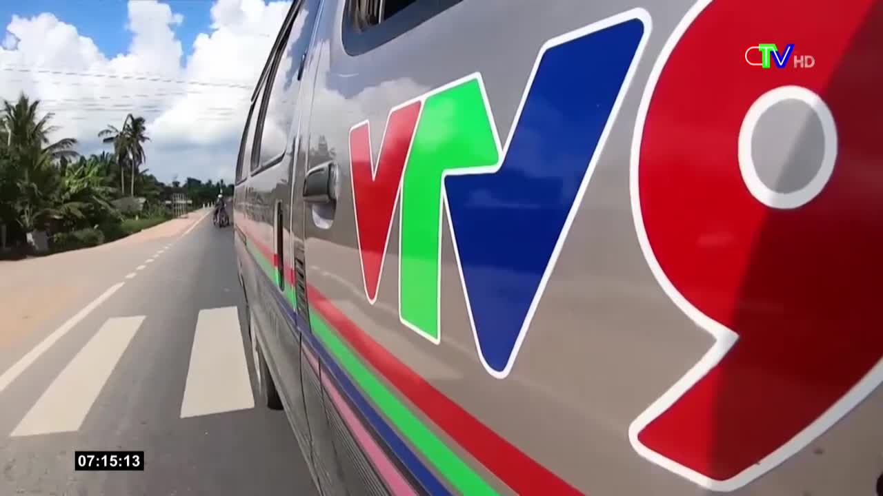 A silver van with the VTV9 logo speeds down a road, the white lines blurring as it passes. Palm trees and other greenery line the road, and the sky is a clear blue.
