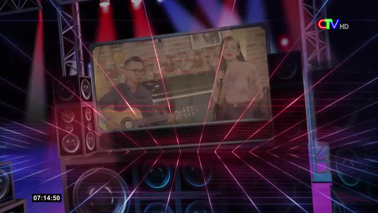 A screen displays a man with glasses playing guitar next to a woman singing into a microphone. Red and blue lights crisscross the stage, while a speaker system sits on either side.
