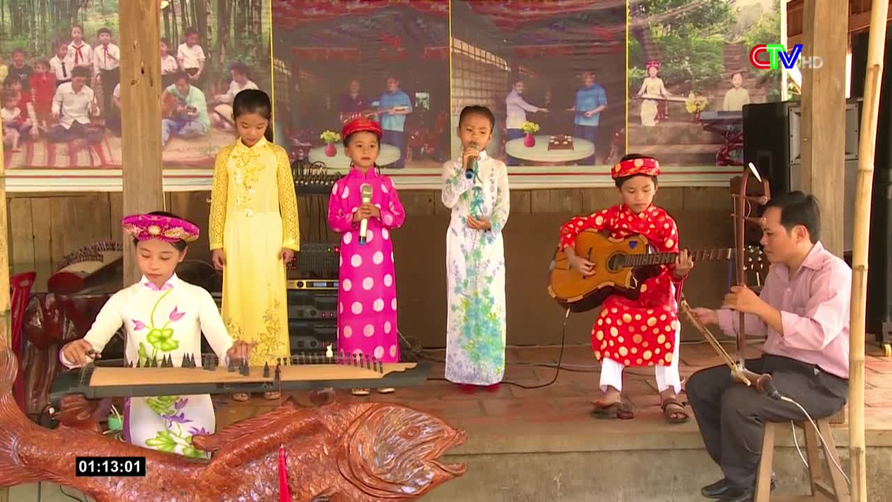 Children in vibrant áo dài dresses and hats perform on a stage, one plucking a stringed instrument, another singing into a microphone. A man plays a bowed instrument alongside a young guitarist, all part of a televised performance from Bac Lieu, Vietnam.

