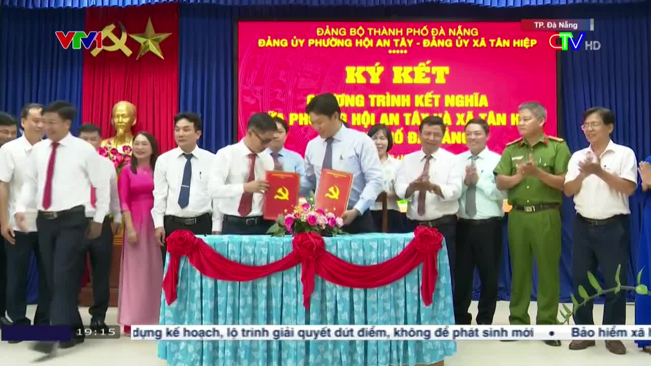 Two men in white shirts and ties are exchanging documents, each bearing a hammer and sickle.  A group of people behind them clap, while the VTV logo and Vietnamese text are visible on the backdrop.
