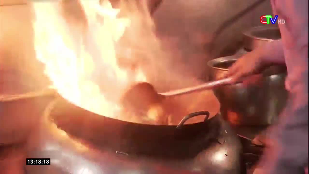 Flames erupt from a wok as a cook stirs the contents with a long-handled spoon. The scene is captured on Bac Lieu TV, a Vietnamese broadcast.
