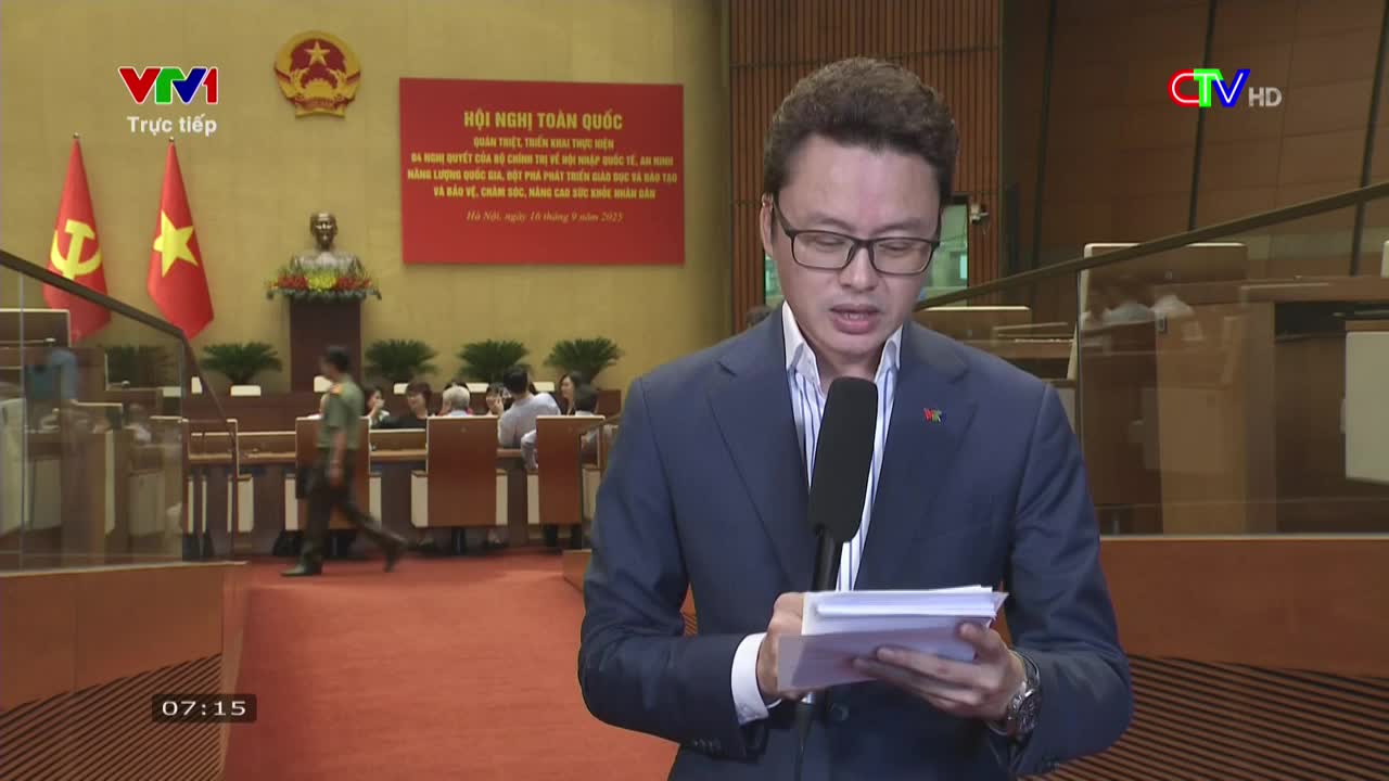 A news anchor in a blue suit speaks into a microphone, reading from a stack of papers. Behind him, a conference is underway, with attendees seated at long tables and flags of Vietnam on display.
