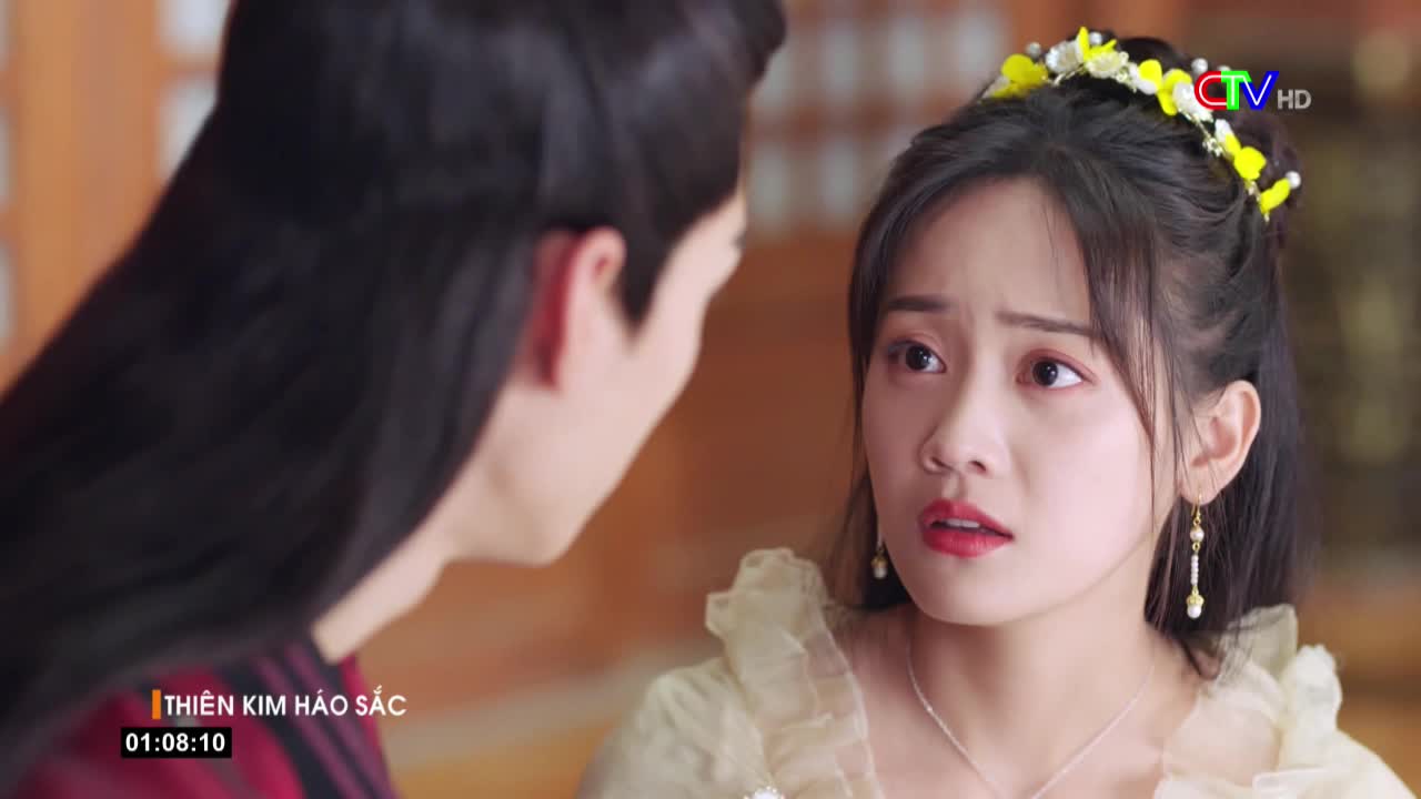 A woman with a floral crown stares, her expression a mix of surprise and concern.  The man facing her is mostly out of frame, but his dark hair and the angle of his face suggest a serious conversation.
