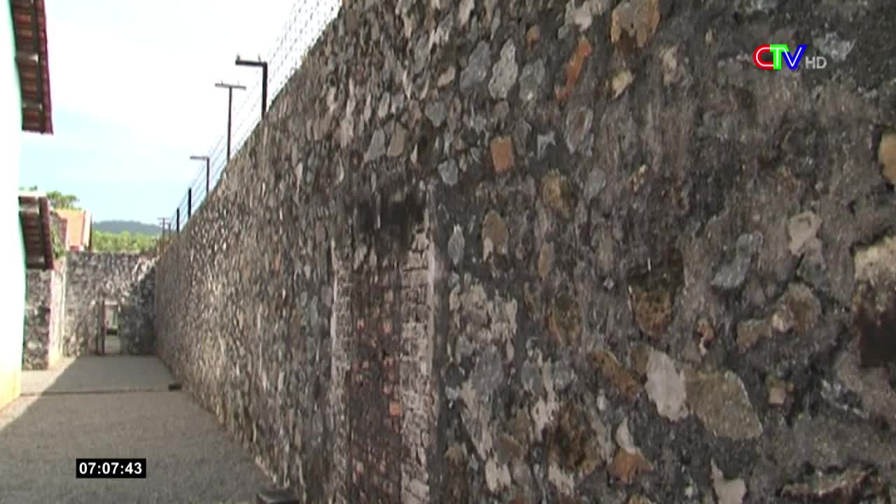 A long, weathered stone wall stretches ahead, topped with barbed wire, and a narrow passage runs alongside it. The sun casts shadows across the path, and the rough texture of the wall is clear.
