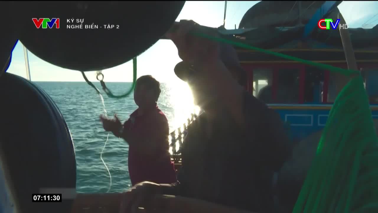 A man in shadow pulls on a green rope, while another man in a pink shirt looks on. The scene plays out on a boat, with the sun reflecting off the water.
A man in shadow pulls on a green rope, while another man in a pink shirt looks on. The scene plays out on a boat, with the sun reflecting off the water.