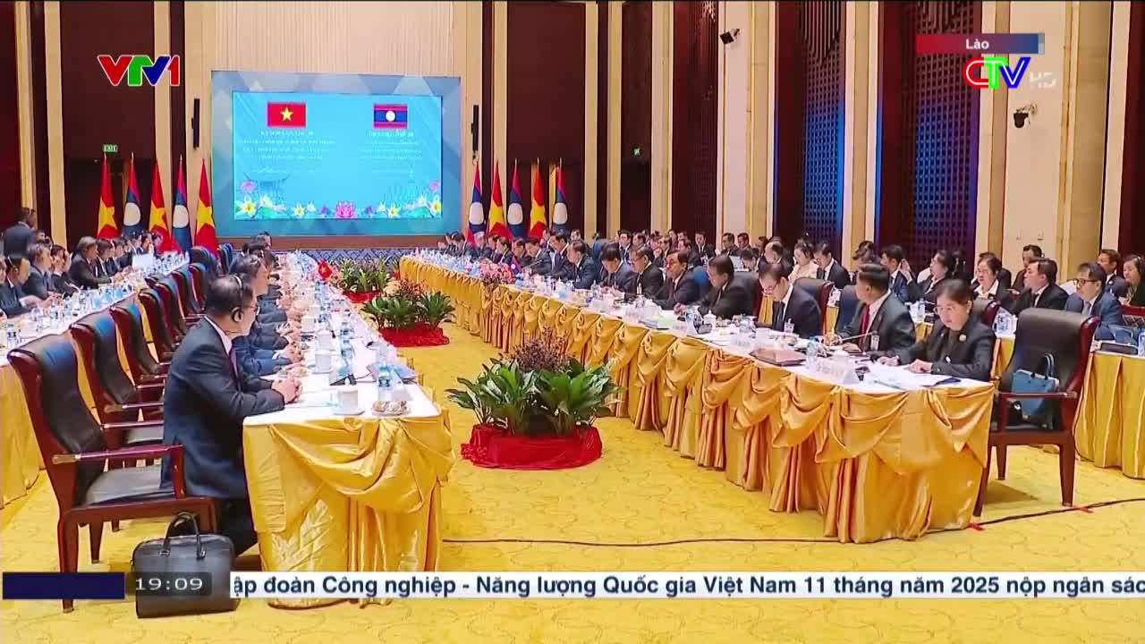 A long table stretches across the room, lined with people in suits, facing a screen displaying flags. The event is being broadcast on Lao TV, with a VTV logo visible in the corner.
A long table stretches across the room, lined with people in suits, facing a screen displaying flags. The event is being broadcast on Lao TV, with a VTV logo visible in the corner.