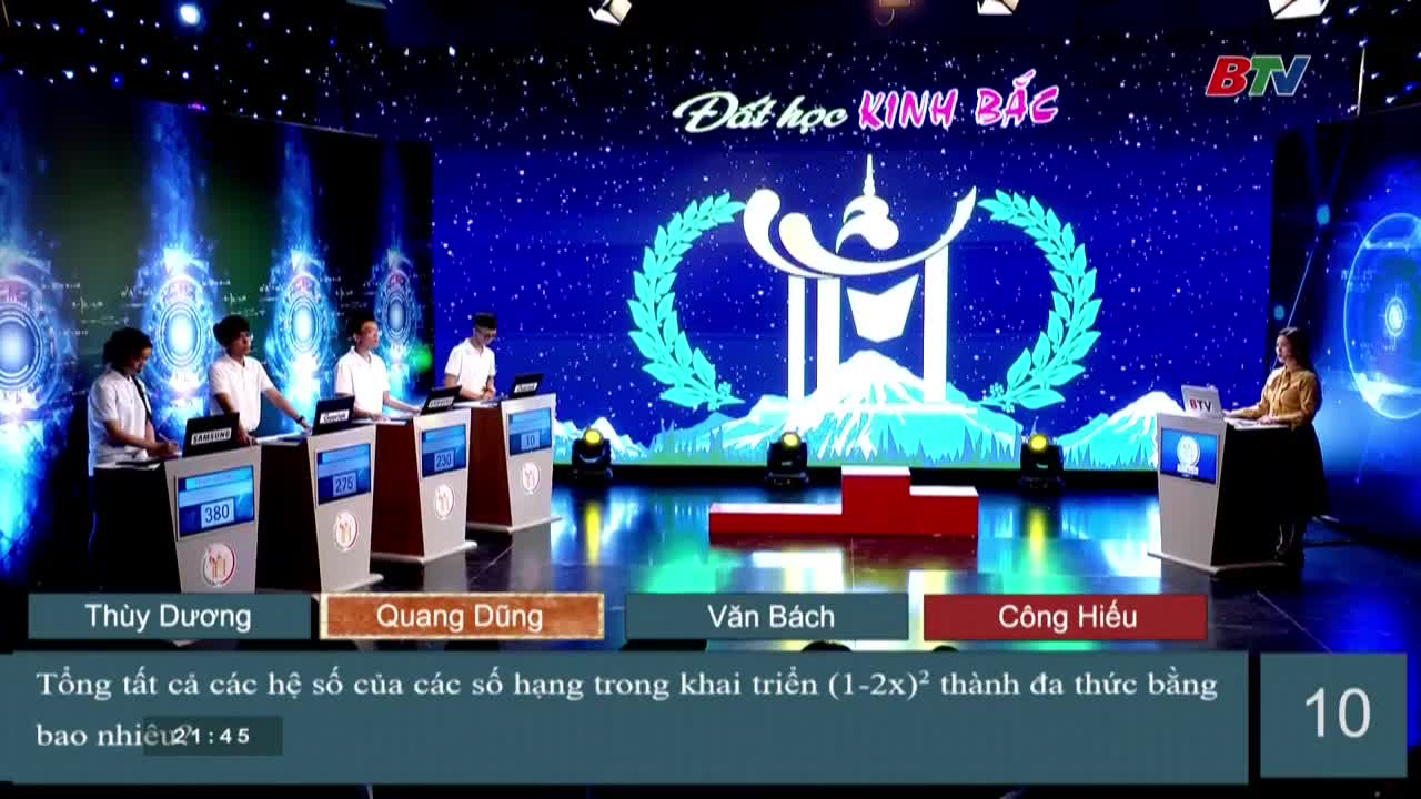 Four young men stand at podiums, each with a lit-up display showing a number. A woman in a yellow shirt stands at a lectern, the countdown clock ticking down on the side.
