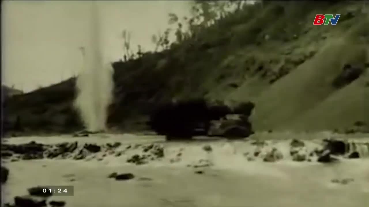 A geyser erupts, shooting a column of water high into the air. A dark vehicle sits on the road below, near the edge of the water.

