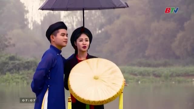 Under a black umbrella, a man and woman in traditional Vietnamese clothing stand together, the woman holding a large, round drum. The scene, likely from a Bac Ninh TV broadcast, unfolds near a misty body of water.
