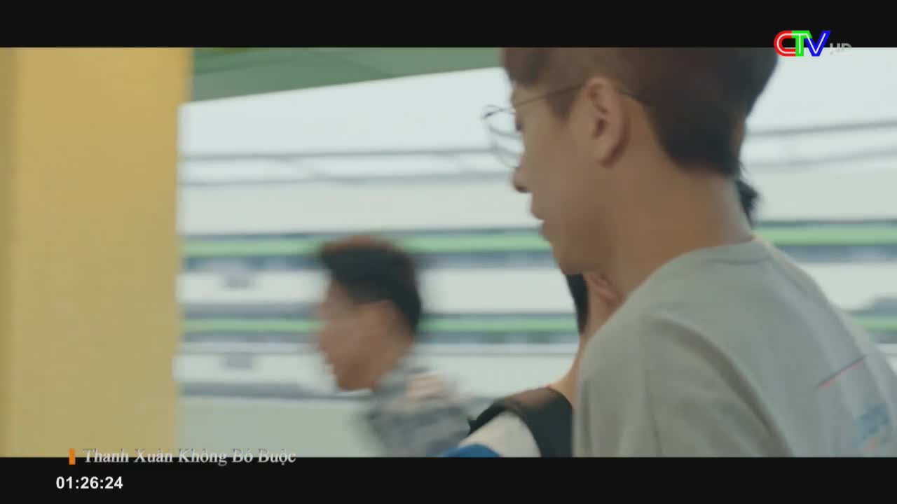 A young man in glasses turns his head, looking forward as he walks. Another person blurs past him in the background, moving quickly.
