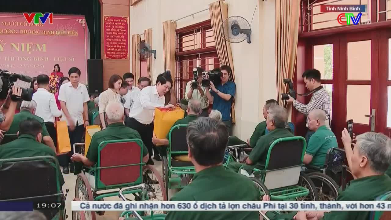 A group of men in white shirts and a few women are distributing yellow packages to veterans in wheelchairs. Camera operators from VTV1 and Ca Mau TV film the event.

