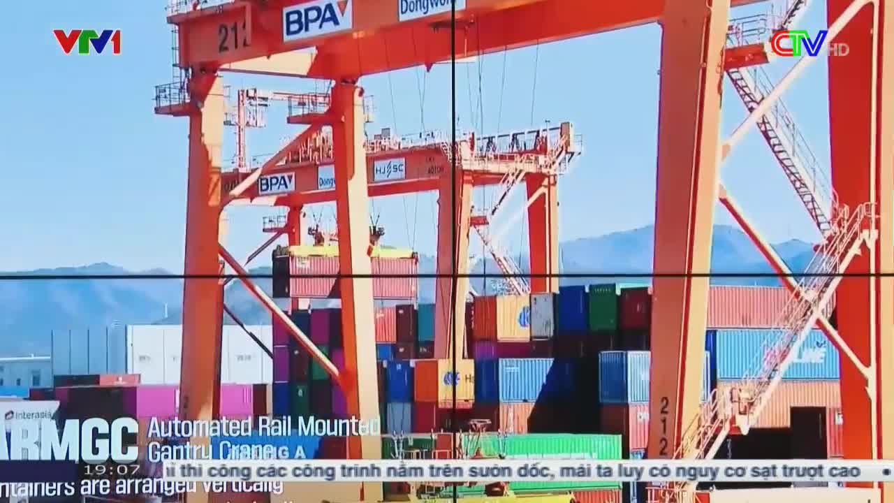 A massive orange crane is lifting a shipping container, its shadow falling across a stack of colorful boxes. The scene is broadcast on Ca Mau TV, with the VTV1 logo in the corner.

