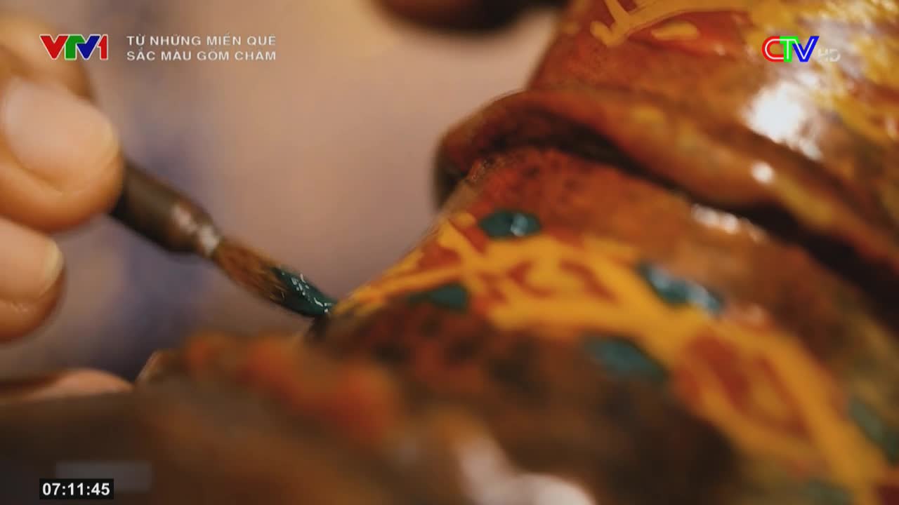 A hand carefully applies a dab of teal paint to a detailed, patterned surface.  The scene is being broadcast on Ca Mau TV, as the VTV logo and Vietnamese script indicate.
