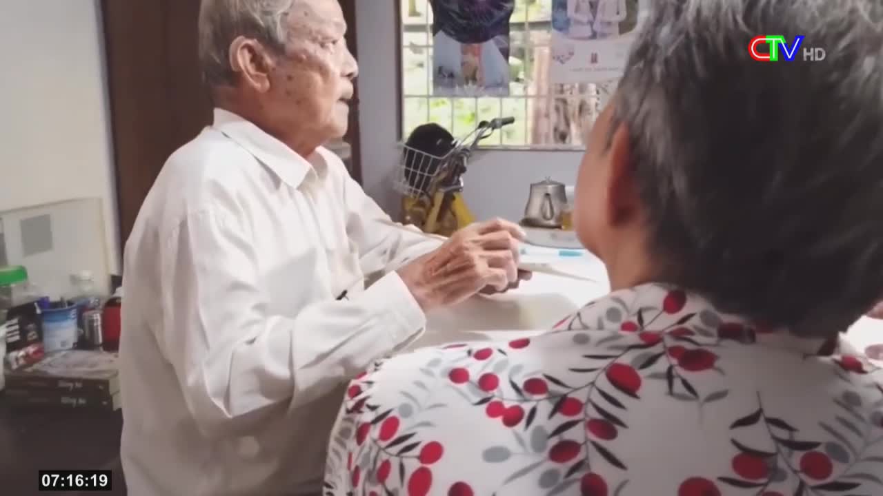 An older man in a white shirt holds a small object out to a person wearing a patterned shirt. The Ca Mau TV logo is visible in the top right corner.
