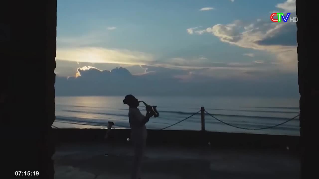 Framed by the dark stone, a figure stands silhouetted, playing a saxophone against the backdrop of a calm sea. The sky is painted with soft hues as the sun begins to rise, and the waves gently lap the shore.
