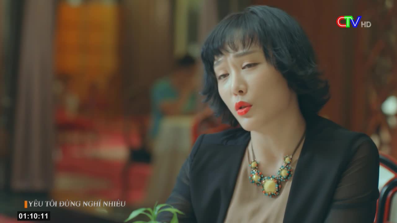 A woman with red lipstick and a jeweled necklace sits, her expression troubled, as the Ca Mau TV logo appears in the corner. The time on the screen reads 01:10:11, and the Vietnamese title suggests a dramatic scene.
