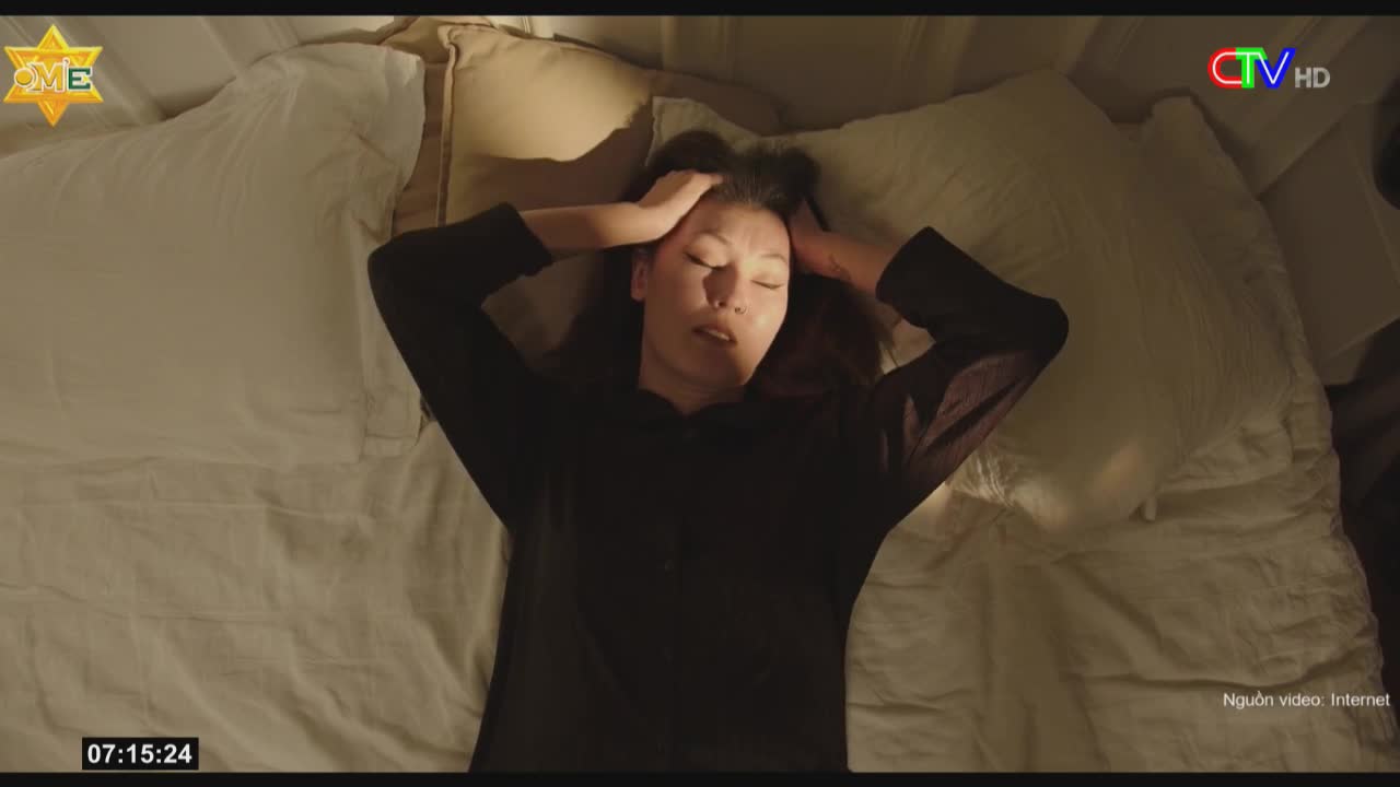A woman, lying on a bed, presses her hands against her temples. Her eyes are closed, and her expression suggests distress.
