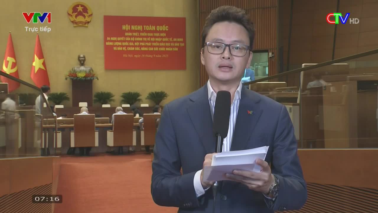 A man in a suit, holding a microphone and papers, addresses the camera in a large hall, likely for a broadcast on VTV1. The Ca Mau TV logo is visible, and a banner in Vietnamese hangs behind him.
