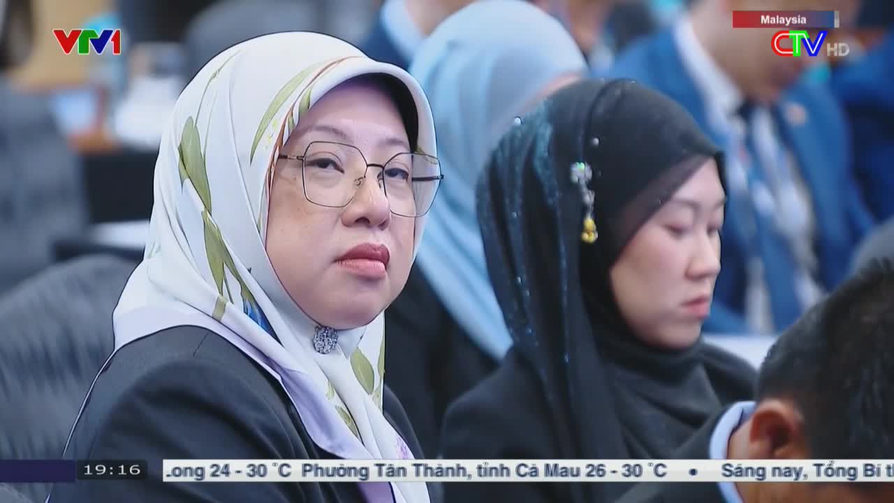 A woman in a patterned headscarf and glasses stares forward, her expression serious. The Ca Mau TV broadcast shows a conference, with a Malaysian flag logo in the corner.
