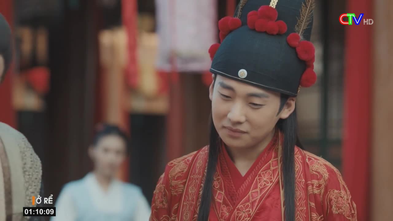 A young man, dressed in elaborate red and gold robes, looks down pensively. The scene is from a Ca Mau TV broadcast, with the show's title and time displayed in the corner.
