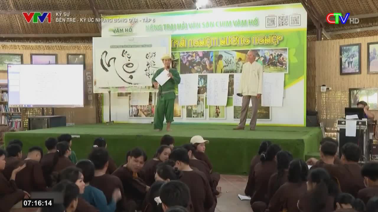 A man in green overalls speaks into a microphone on a stage, while another man stands beside him. The audience, seated on the floor, listens to the presentation, which is likely being broadcast on Ca Mau TV.
