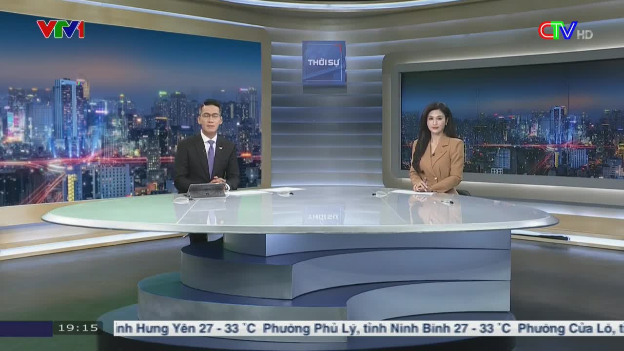 Two news anchors sit at a large, curved desk, facing the camera. Behind them, a cityscape glows on the screens, the backdrop for the evening news broadcast from Vietnam.
