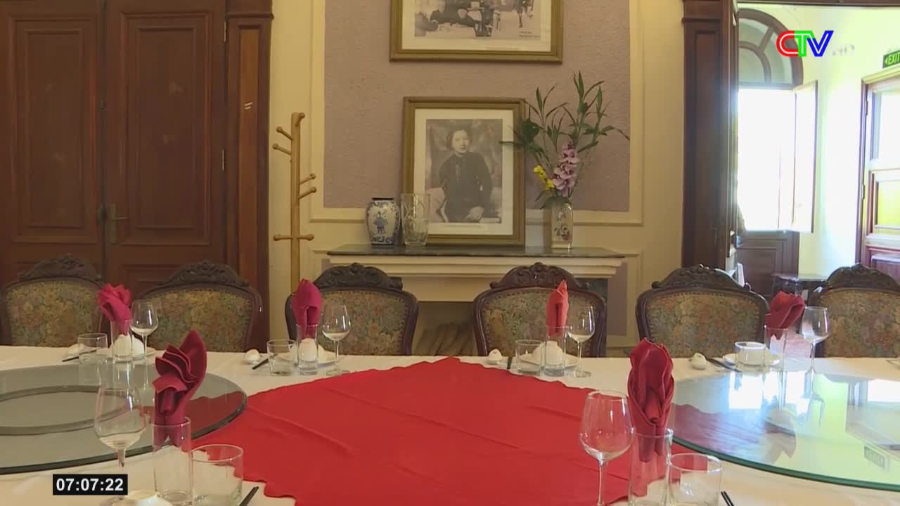 A long table is set for a formal meal, with red napkins and crystal glasses reflecting the light. The Ca Mau TV logo is visible in the corner, and a framed portrait hangs on the wall behind the table.
