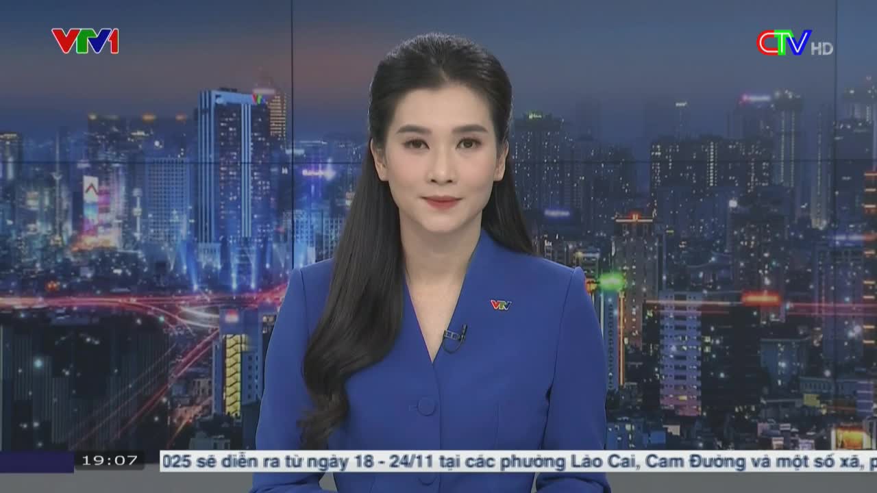 A woman in a blue blazer stands in front of a cityscape backdrop, speaking on Vietnam's Ca Mau TV. The time is 19:07, and text scrolls at the bottom of the screen.
