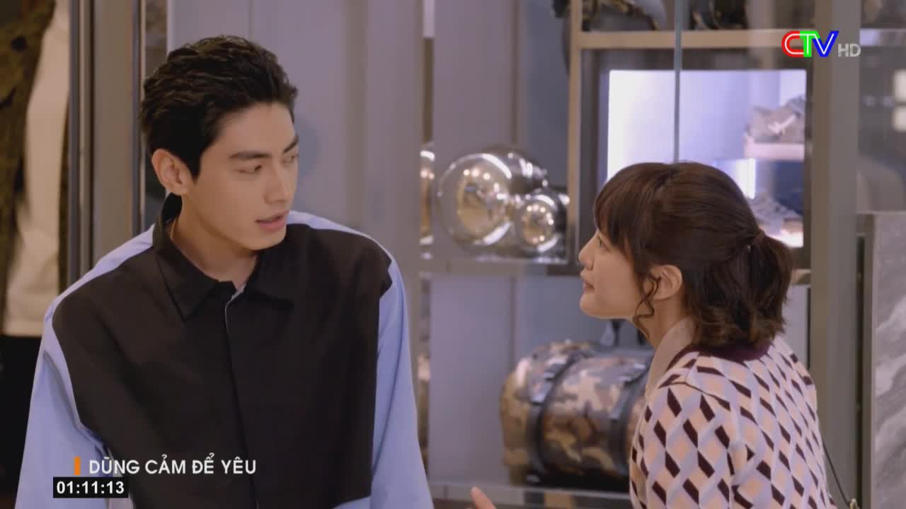 The man in the black and blue shirt looks at the woman, listening intently. She gestures with her hand, her expression serious as she speaks.
