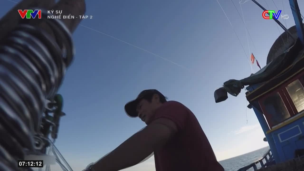 A man, wearing a cap and red shirt, leans over the side of a boat, the Ca Mau TV logo visible in the corner. The bright sky and ocean stretch out behind him as he works.
