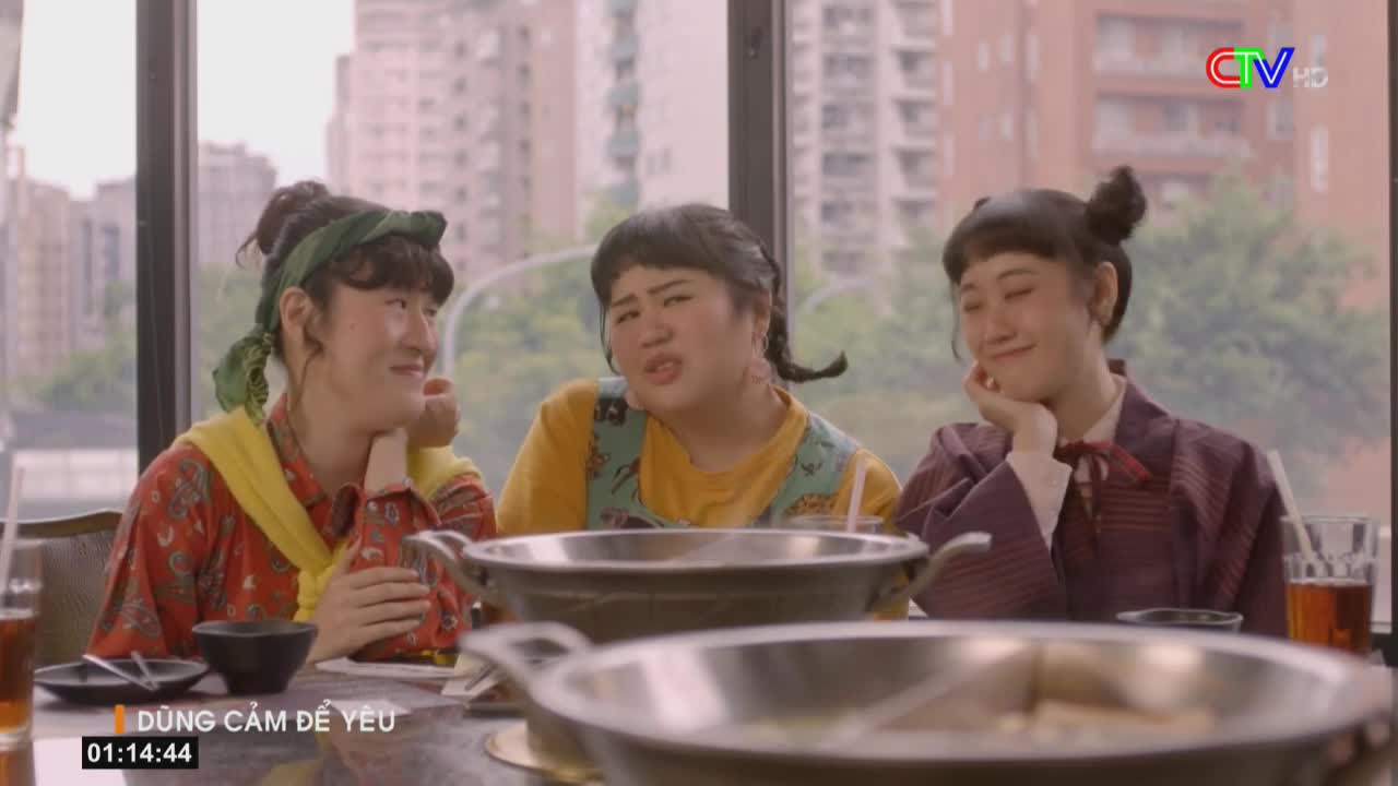 Three women are seated around a table, each with a hand propping up their chin. A steaming pot sits between them, and they seem to be in the middle of a conversation, as the middle woman is speaking.
