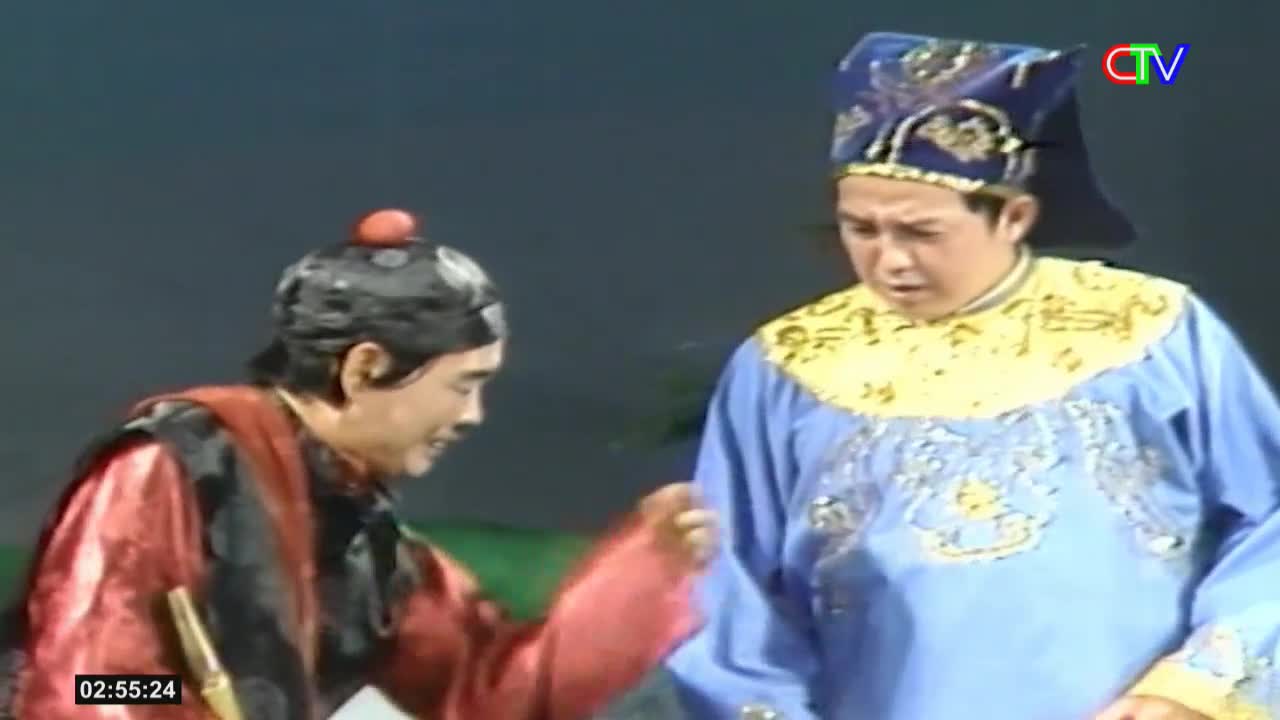 A man in a red robe gestures emphatically towards another man in a bright blue, ornate costume. The scene unfolds on a stage, likely part of a performance broadcast by Ca Mau TV.