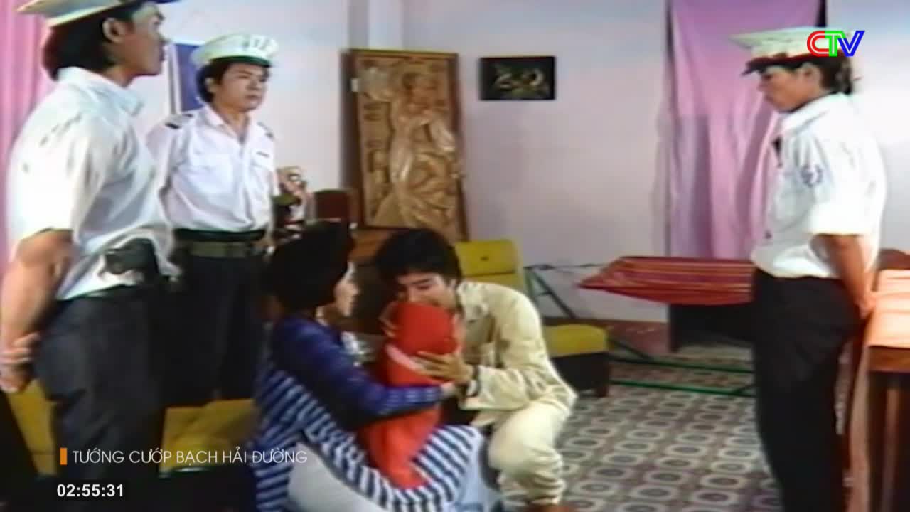 Two men in uniform stand watch as a man embraces a woman holding a red bundle. The scene unfolds indoors, likely a domestic setting, with a hint of official presence from the men's attire.