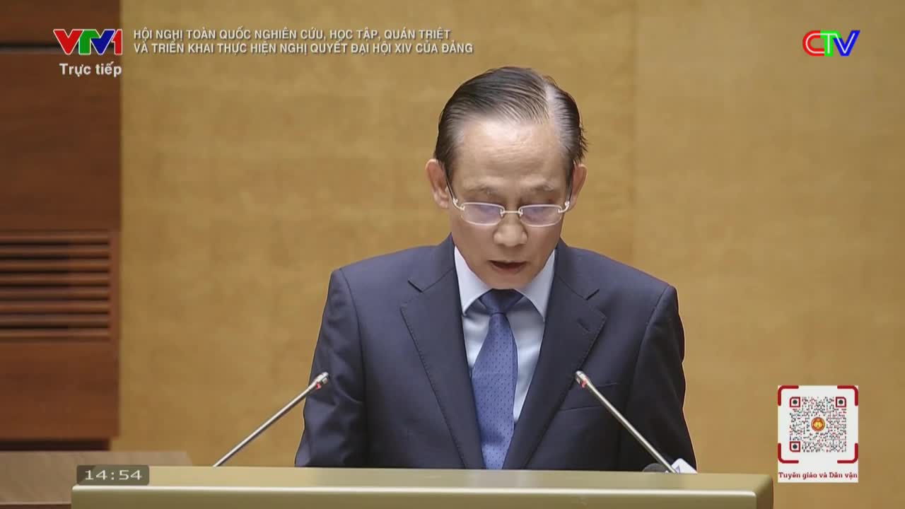 A man in a dark suit and blue tie stands at a podium, speaking into two microphones. The screen displays "Trực tiếp" and the VTV logo, indicating a live broadcast from Vietnam.