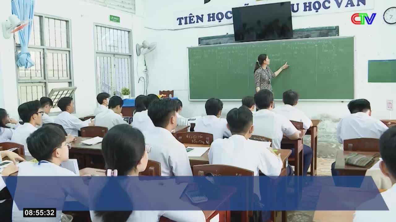 A teacher stands at the blackboard, her arm extended, pointing to equations. Students in white shirts sit at their desks, facing forward.