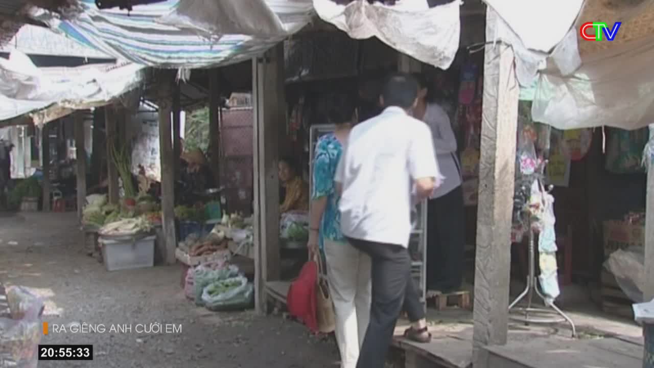 A man in a white shirt walks past a stall in this Vietnamese market.  He's heading towards a woman in a blue and white patterned top, carrying a red bag.