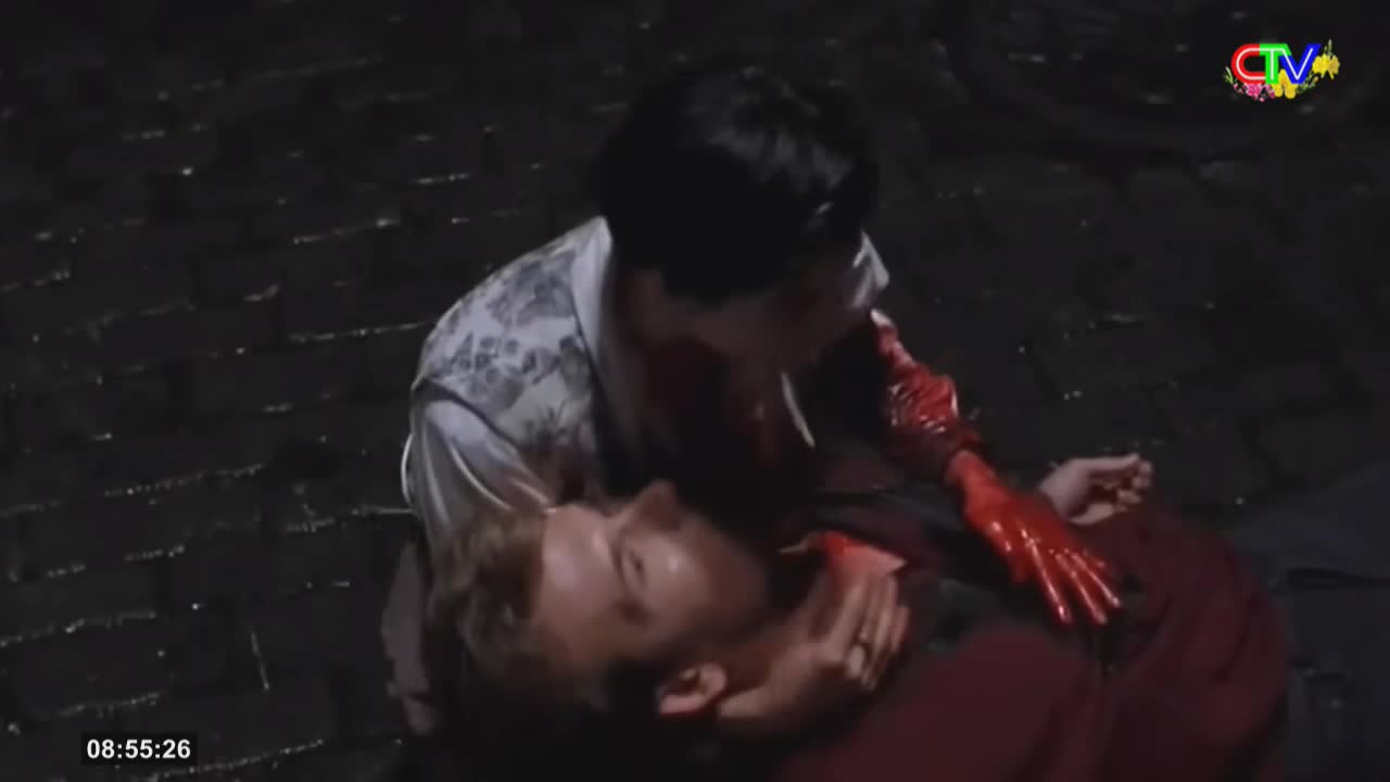 A figure in a patterned shirt leans over another person lying on the wet cobblestones, their hand covered in bright red liquid. The scene is lit by a dim, overhead light, casting long shadows.
