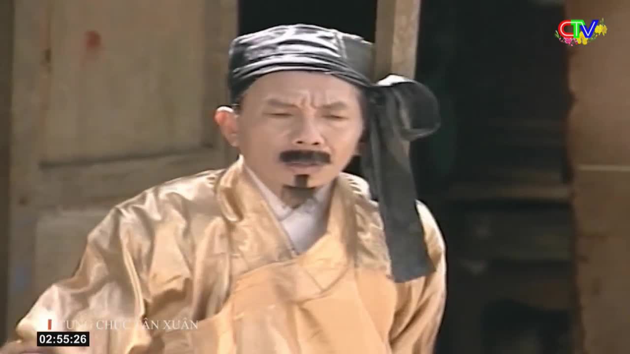 A man in traditional Vietnamese attire, a golden robe and a black cap, stands with a stern expression. He's likely on a set for a Ca Mau TV production, given the logo in the corner.