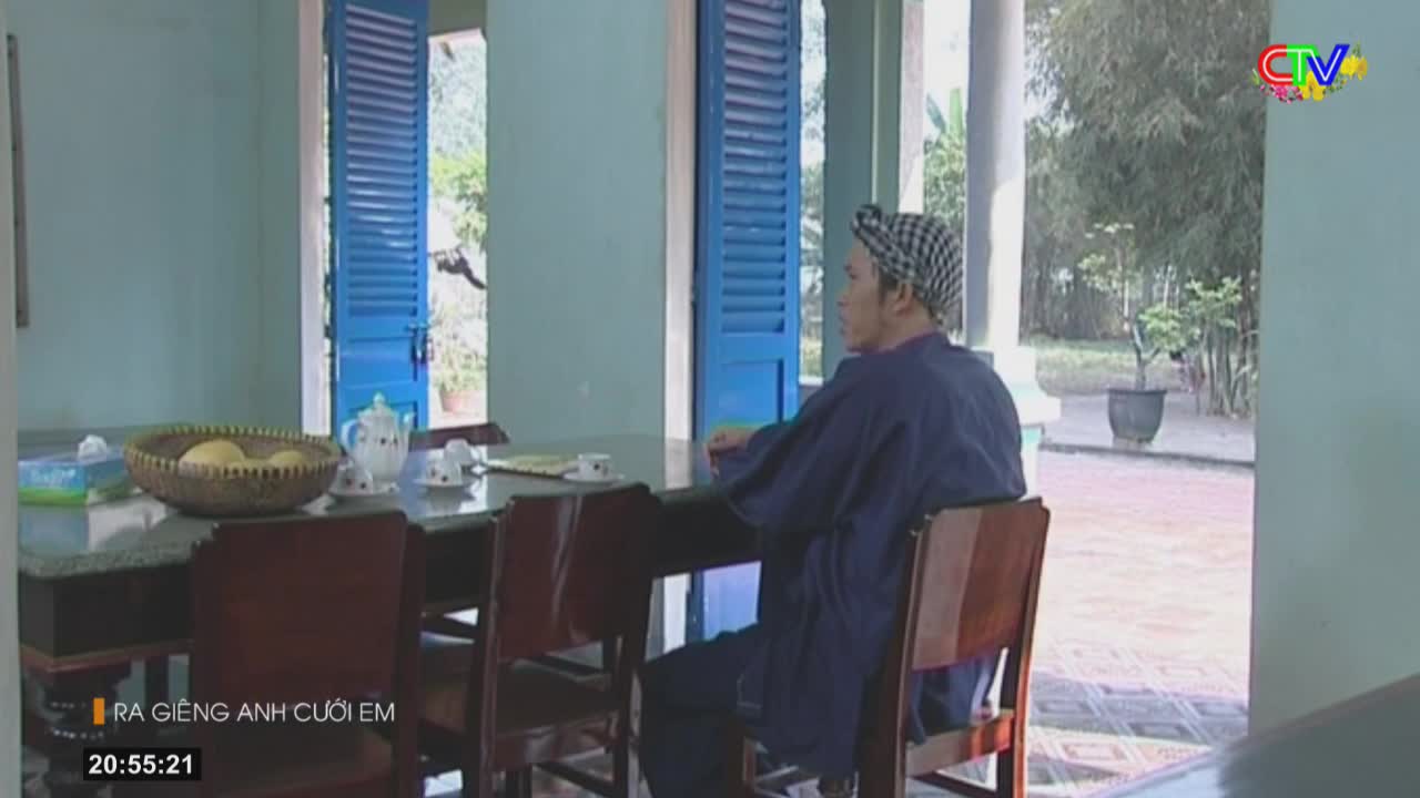 A man in a dark blue robe and checkered headscarf sits at a table, looking out an open doorway. Sunlight streams in, illuminating a fruit bowl and teacups on the table.