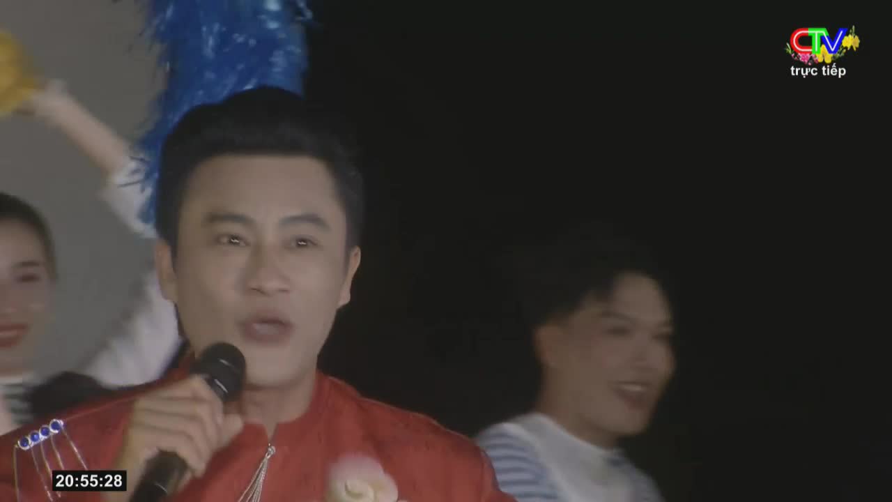A man in a red jacket sings into a microphone, while behind him, two people hold up pom-poms, one yellow and one blue. The Ca Mau TV logo appears in the top right corner.
