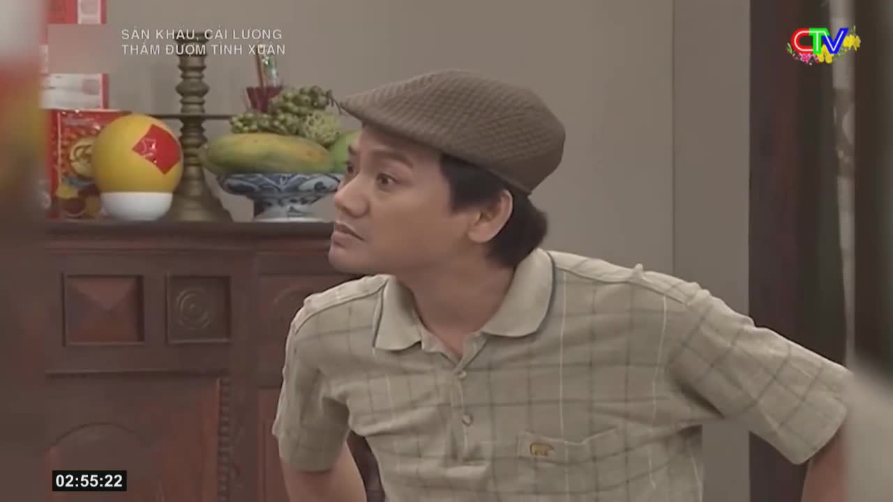 A man in a checkered shirt and flat cap looks off to the side, his expression intent. Behind him, a shelf holds a yellow ball with a red emblem and a bowl of fruit.