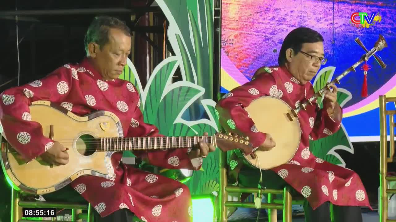 Two men in traditional Vietnamese áo dài are playing instruments on a stage. The man on the left strums an acoustic guitar, while the man on the right plays a lute.