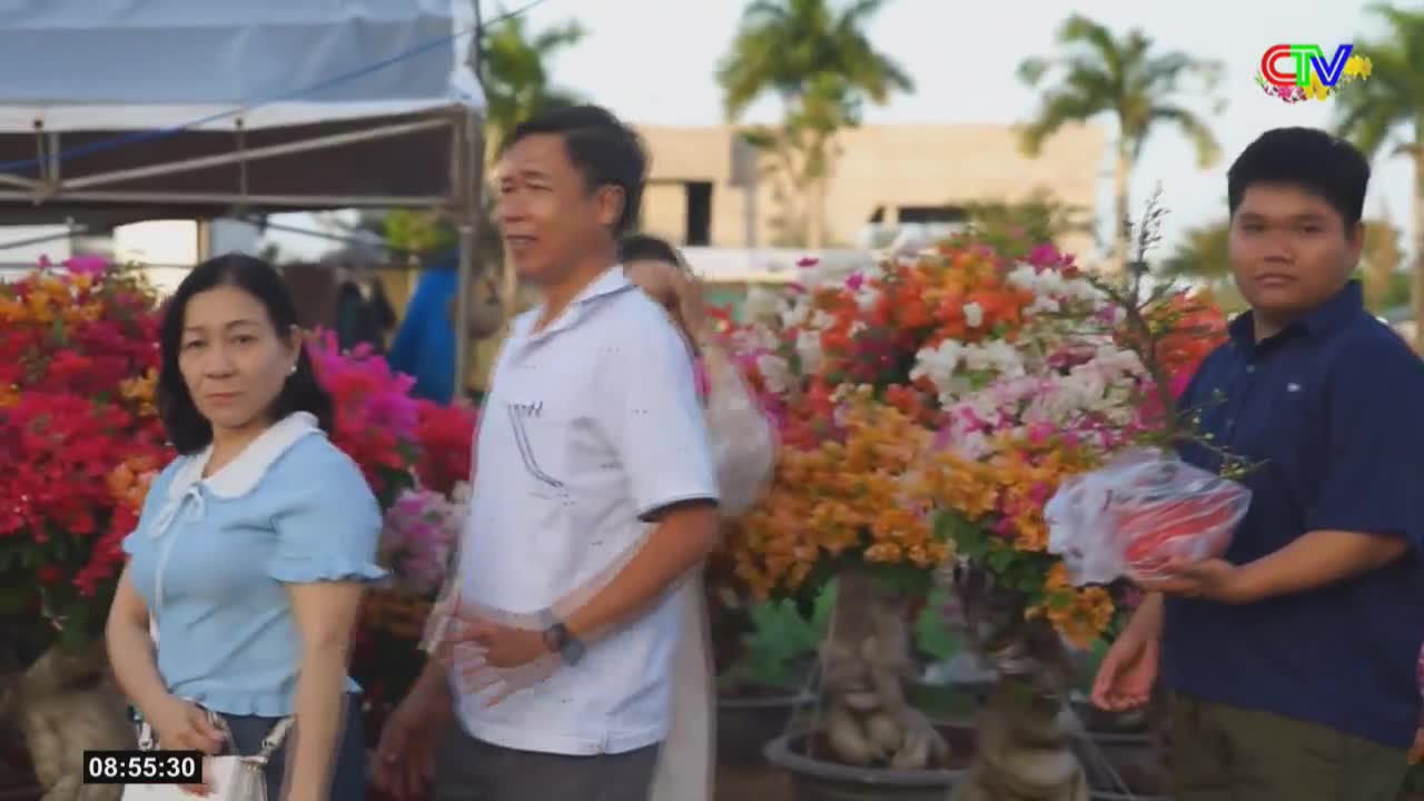 A man in a white polo shirt stands beside a woman in a blue blouse, both looking towards the camera. Behind them, a vibrant display of bougainvillea flowers bursts with color, and a young man holds a small potted plant. The scene unfolds under a bright sky, with palm trees visible in the distance.