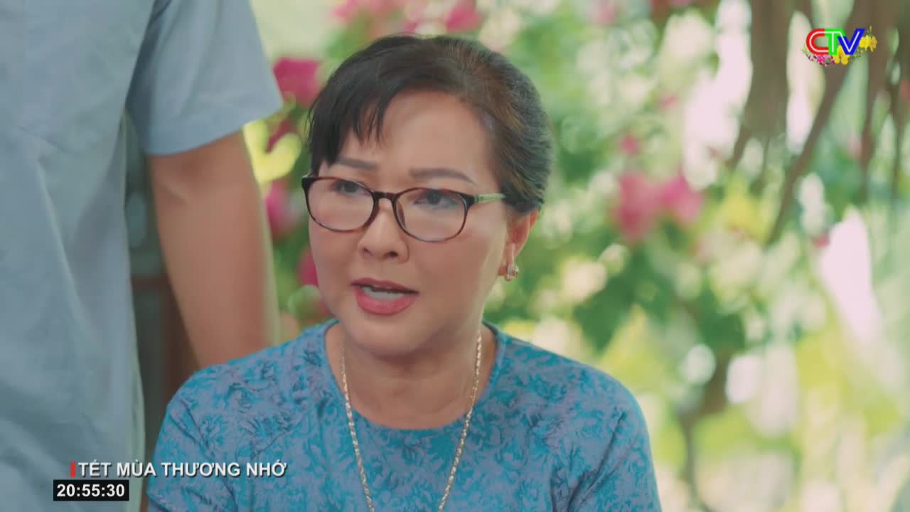 A woman with glasses speaks, her patterned blue shirt visible. Behind her, a person in a light blue shirt stands, and soft pink flowers bloom in the background.