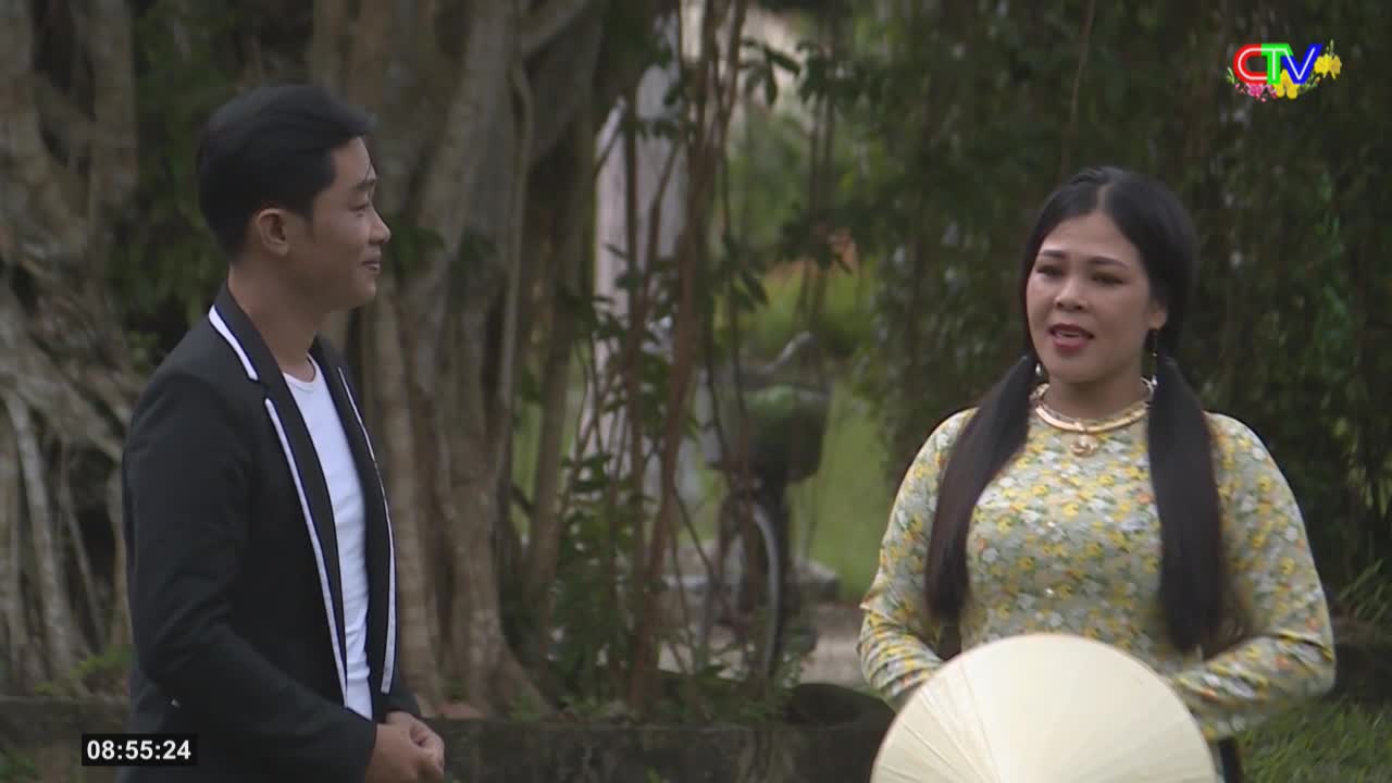 A man in a black jacket with white trim stands to the left, facing a woman in a floral dress. The woman, with her hair in pigtails, holds a conical hat in front of her.