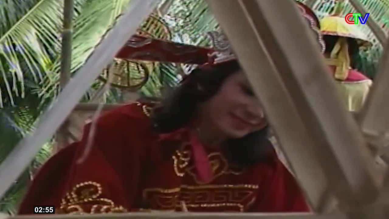 A woman in a red, ornate robe with gold trim smiles as she peeks through bamboo poles. The Ca Mau TV logo is visible in the upper right corner.