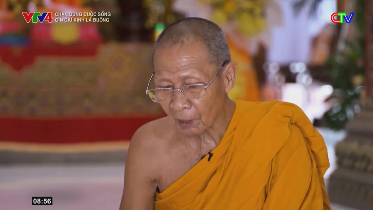 An elderly monk in an orange robe speaks, his glasses perched on his nose. The Ca Mau TV logo appears in the corner, indicating this broadcast from Vietnam.