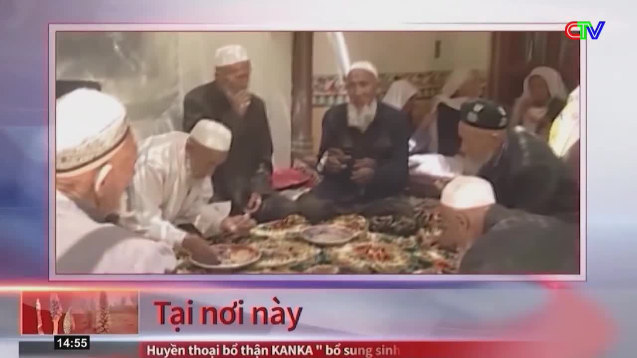Several men in white caps are gathered around a low table, eating from plates of food. One man in a dark jacket is speaking, gesturing with his hand.